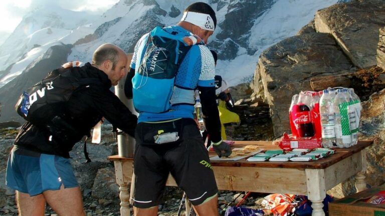 Trail runner refilling soft flasks with water and electrolyte drink at an aid station during a trail race.